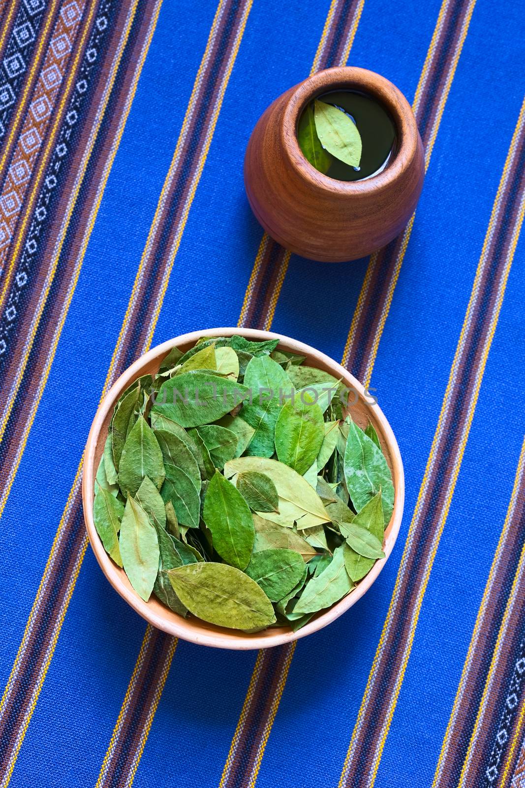 Overhead shot of dried coca leaves in clay bowl with fresh coca tea (mate de coca) on blue fabric, photographed with natural light 