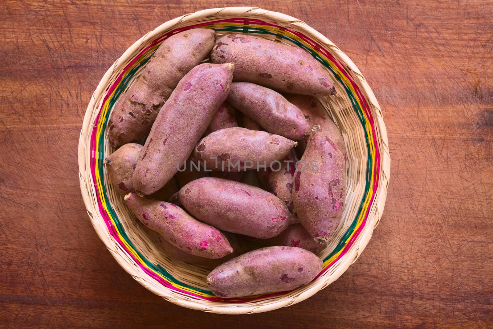 Overhead shot of raw purple sweet potato (lat. Ipomoea batatas) in woven basket on wooden board photographed with natural light   