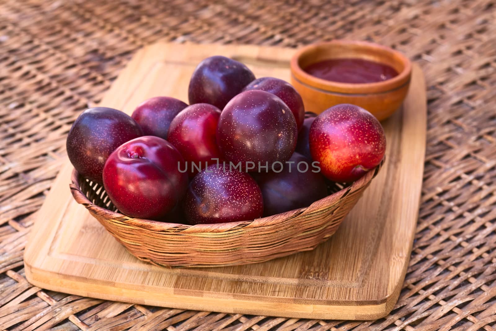 Woven basket filled with satsuma plum with small bowl of plum jam in the back on wooden board photographed with natural light (Selective Focus, Focus on the plums in the front)