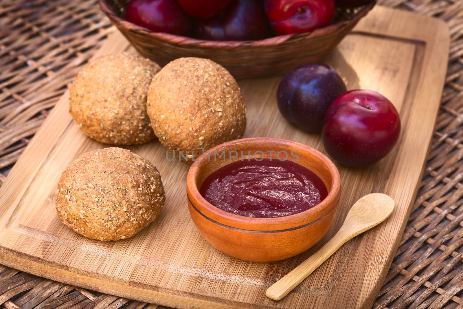 Small wooden bowl of plum jam with wholegrain buns and satsuma plums on wooden board photographed with natural light (Selective Focus, Focus in the middle of the jam and on the front of the bun next to it)