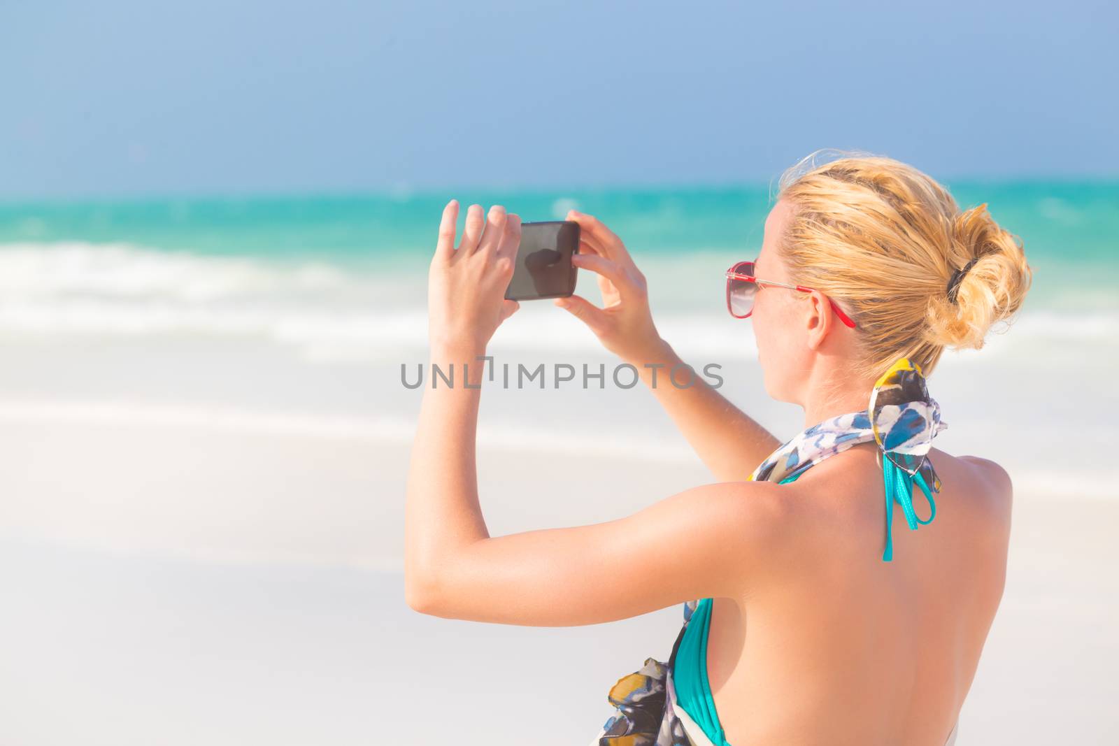 Blonde caucasian woman taking photo of blue tropical beach. Beautiful caucasian model  wearing turquoise swimsuit and colorful scarf on vacations on picture perfect Paje beach, Zanzibar, Tanzania.