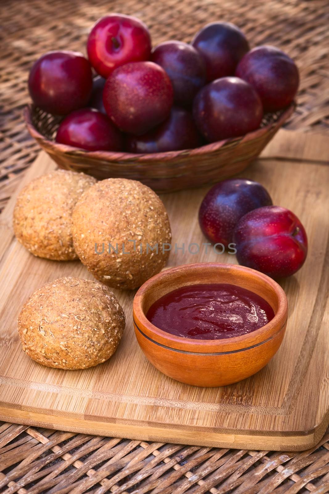 Small wooden bowl of plum jam with wholegrain buns and satsuma plums on wooden board photographed with natural light (Selective Focus, Focus in the middle of the jam and on the front of the bun next to it)