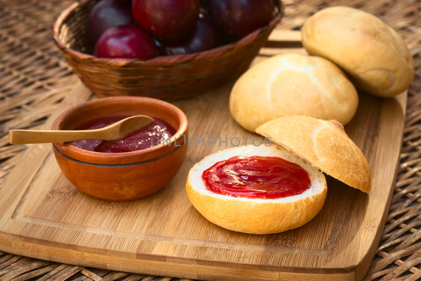 Plum jam spread on bun with plums in basket on wooden board photographed with natural light (Selective Focus, Focus on the front of the jam on the bun)