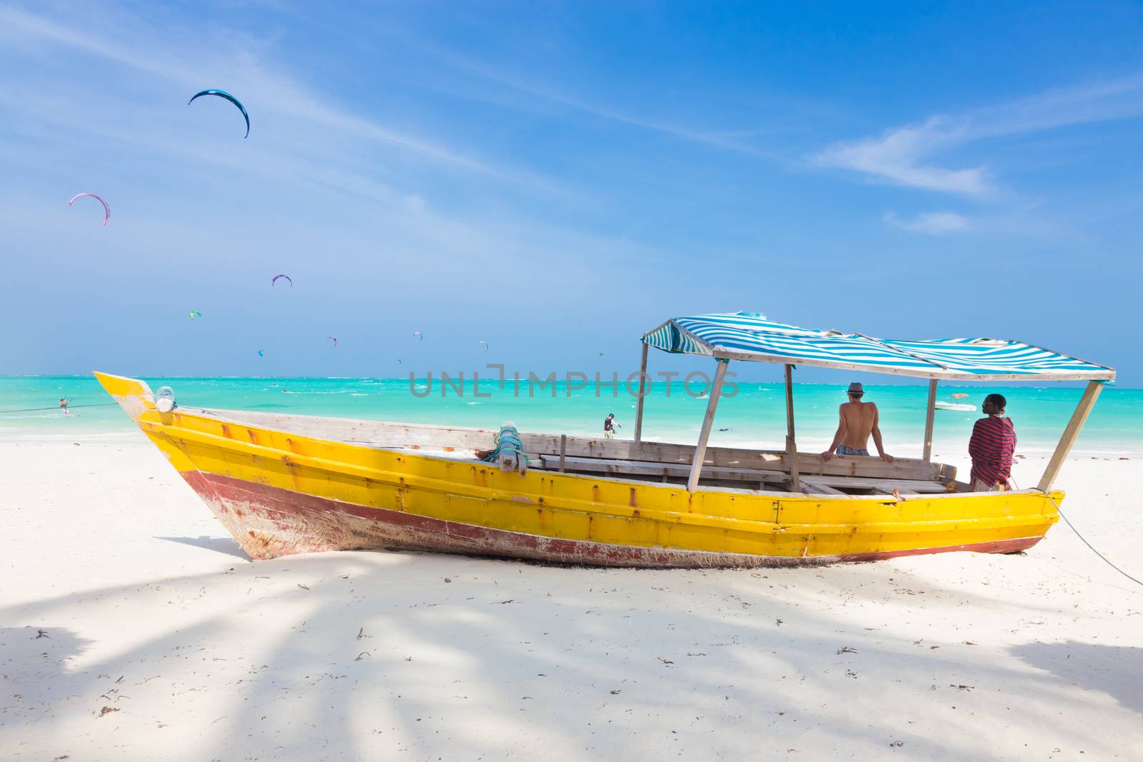Caucasian tourist and Maasai warrior lounging aroundon traditional colorful wooden boat on picture perfect tropical sandy beach on Zanzibar, Tanzania, East Africa. Kiteboarding spot on Paje beach.