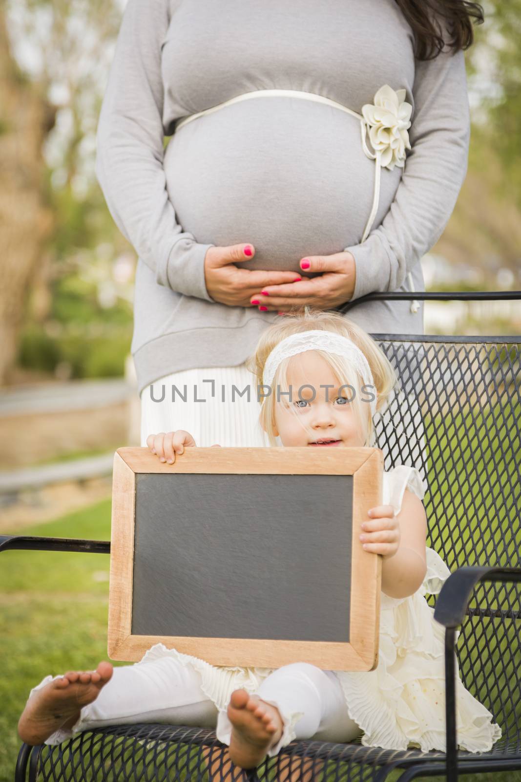 Pregnant Mom Behind Cute Baby Girl Sitting in Chair Holding Blank Blackboard.