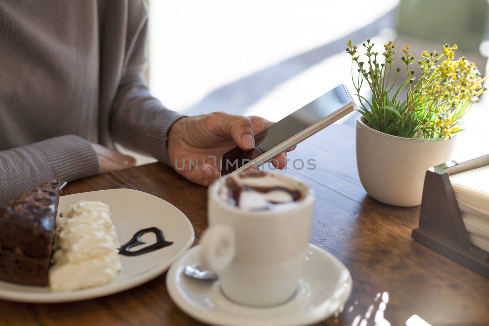 woman hand touch phone next to cake piece and white cup cappuccino coffee with chocolate heart on light brown wooden table