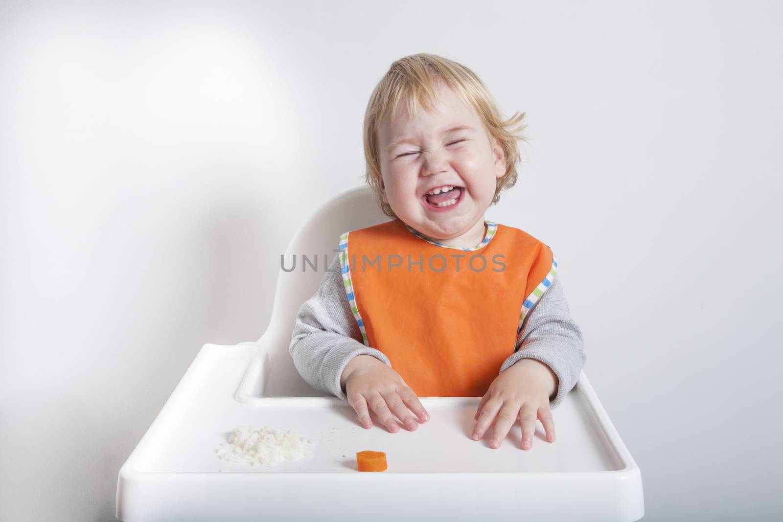 blonde caucasian baby seventeen month age orange bib grey sweater eating rice carrot on white high-chair laughing and smiling