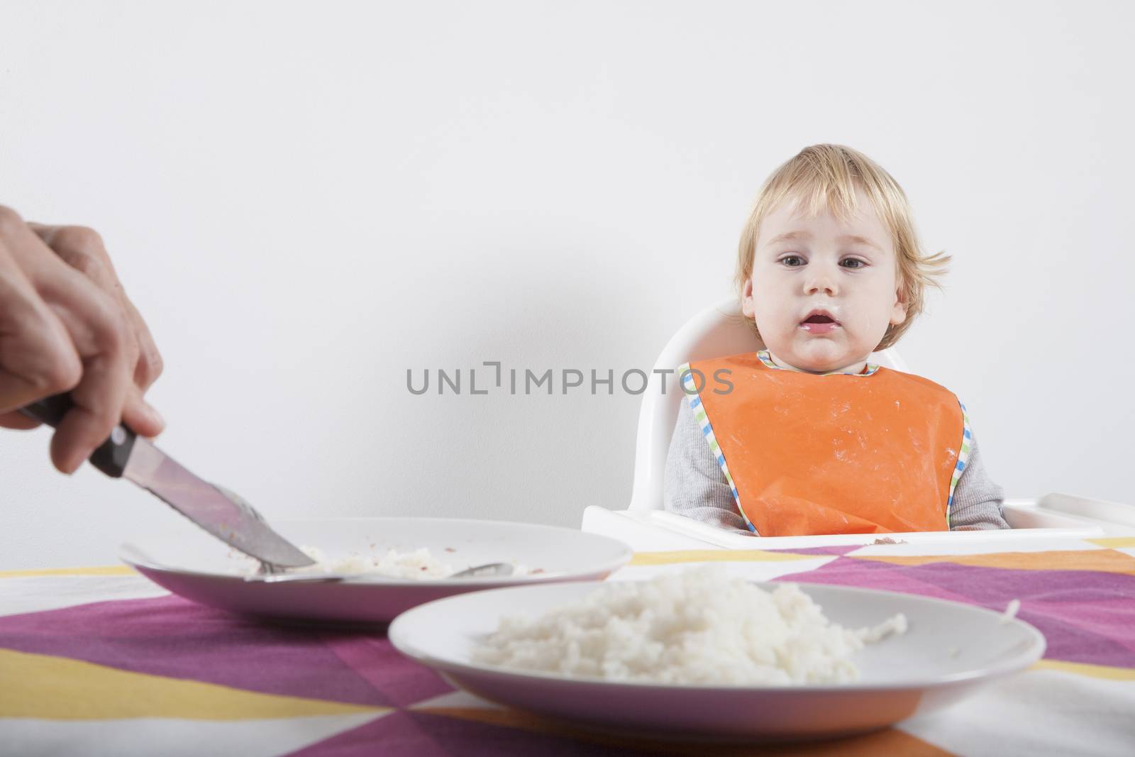 blonde caucasian baby seventeen month age orange bib grey sweater on white high-chair looking at mother hands cutting food with fork on dish