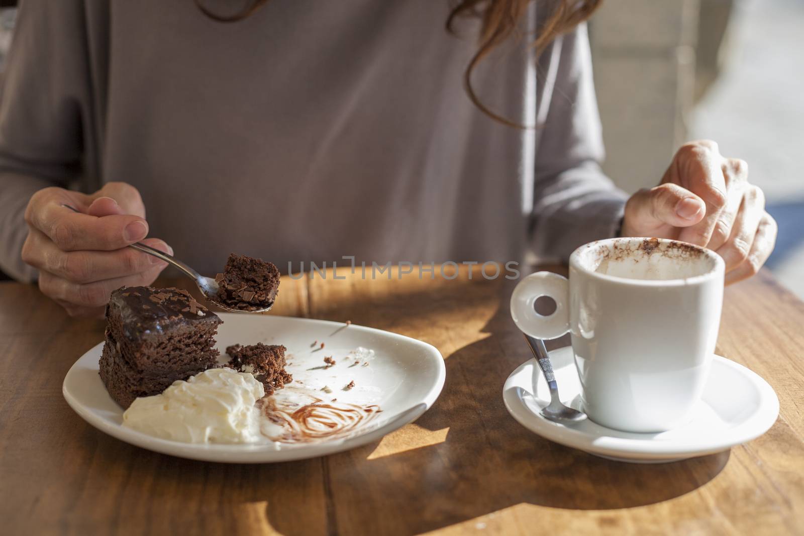 woman hands taking chocolate cake piece with spoon and white small cup cappuccino coffee on light brown wooden table