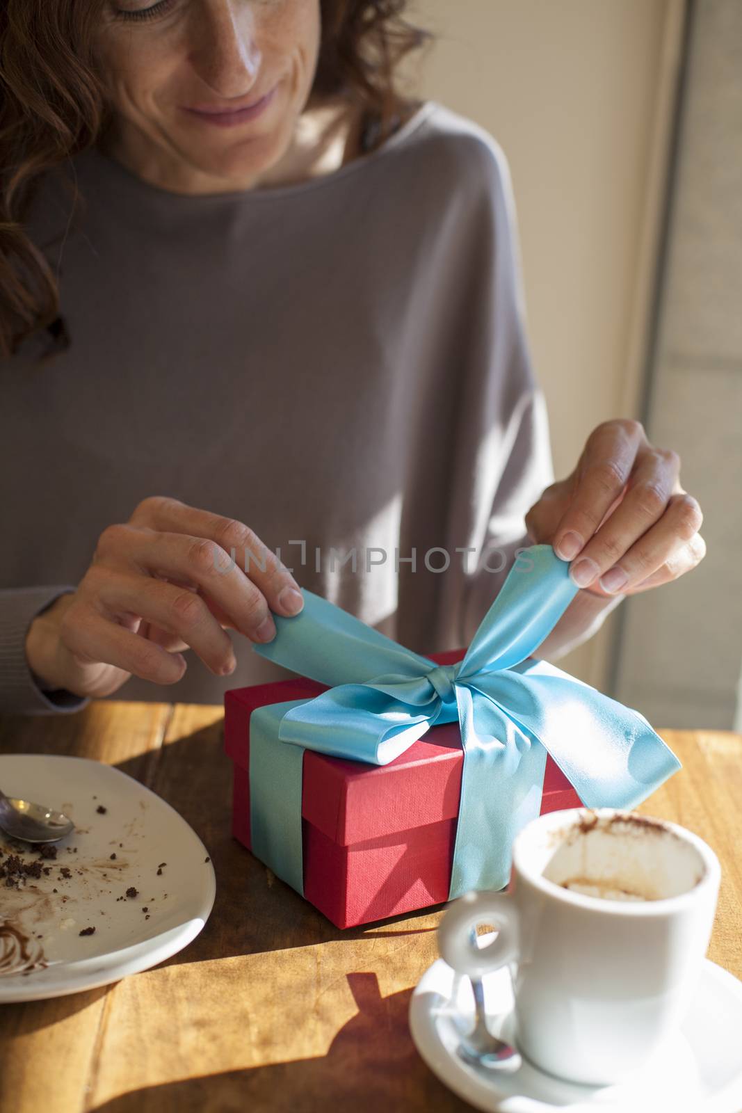 woman open red and blue gift box on light brown wooden table with white small cup cappuccino coffee and chocolate cake dish