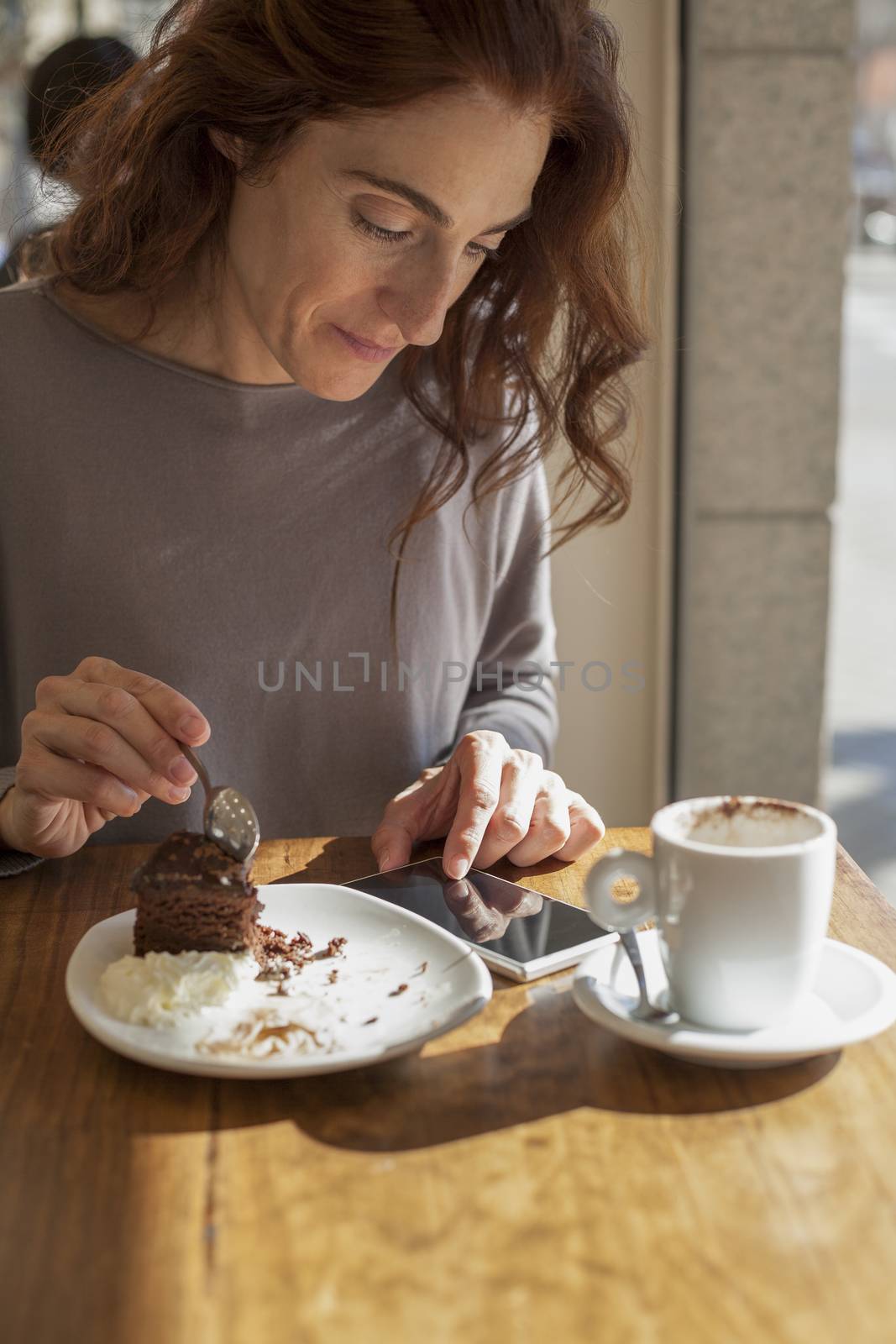 woman taking chocolate cake piece with spoon next to white small cup cappuccino coffee and touching mobile phone blank screen on light brown wooden table