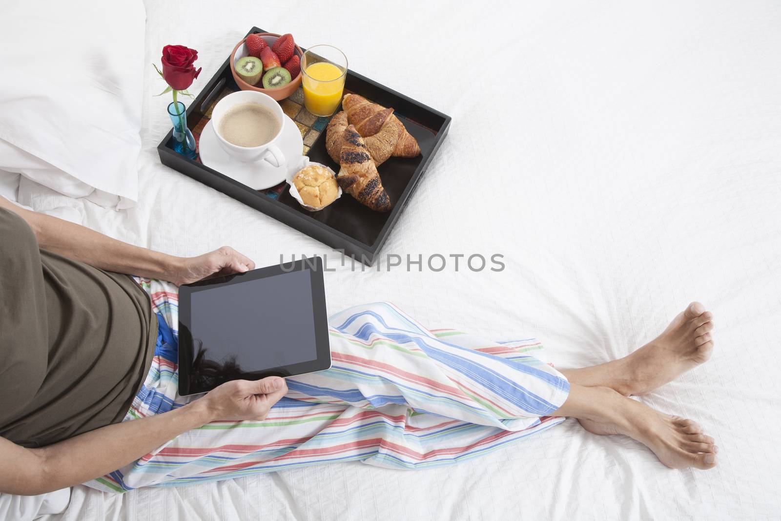 woman green shirt striped pajama pants sitting on white bed with digital tablet blank screen and breakfast tray croissants orange juice strawberry kiwi cupcake red rose flower