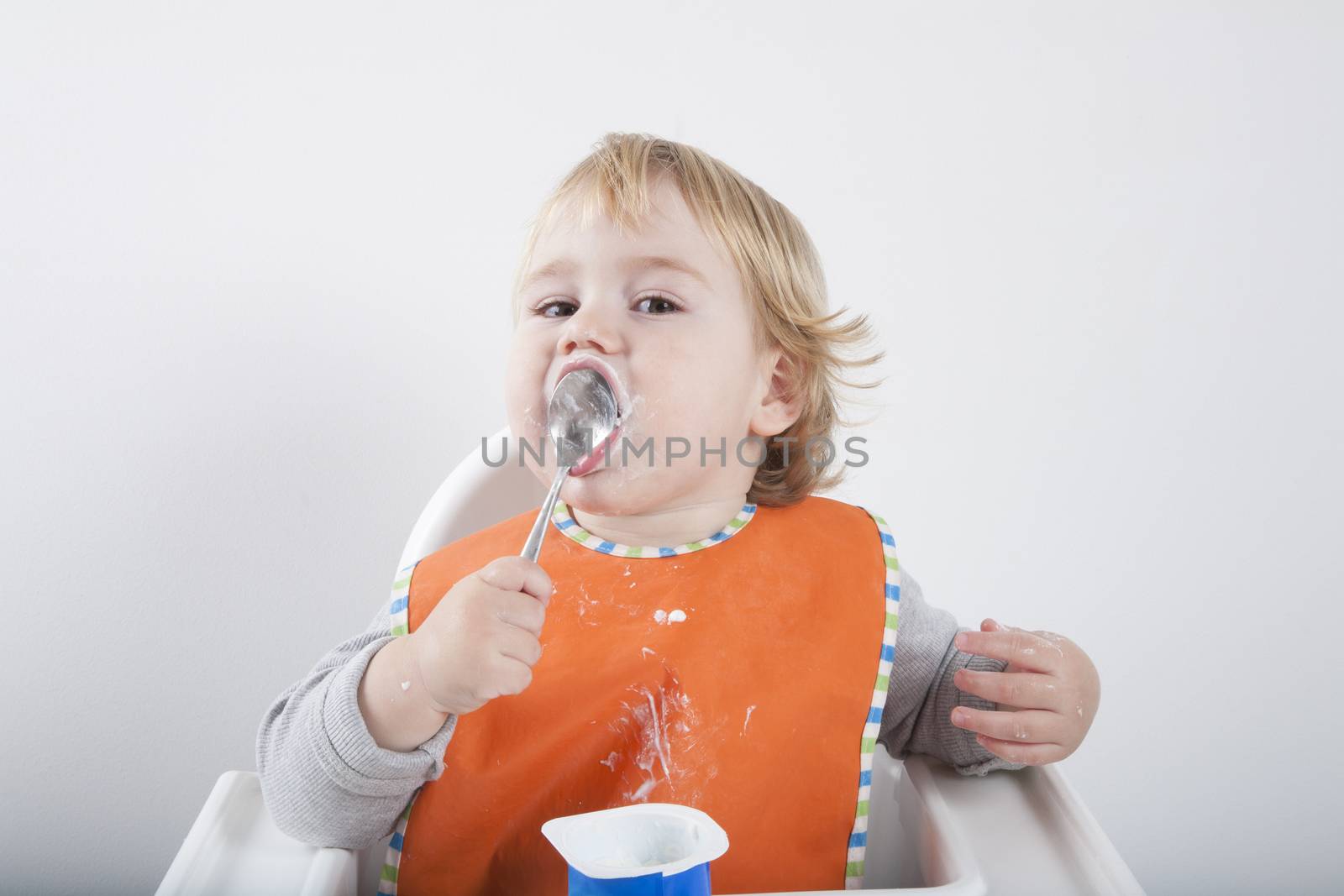 blonde caucasian baby seventeen month age orange bib grey sweater in white high chair licking spoon yogurt