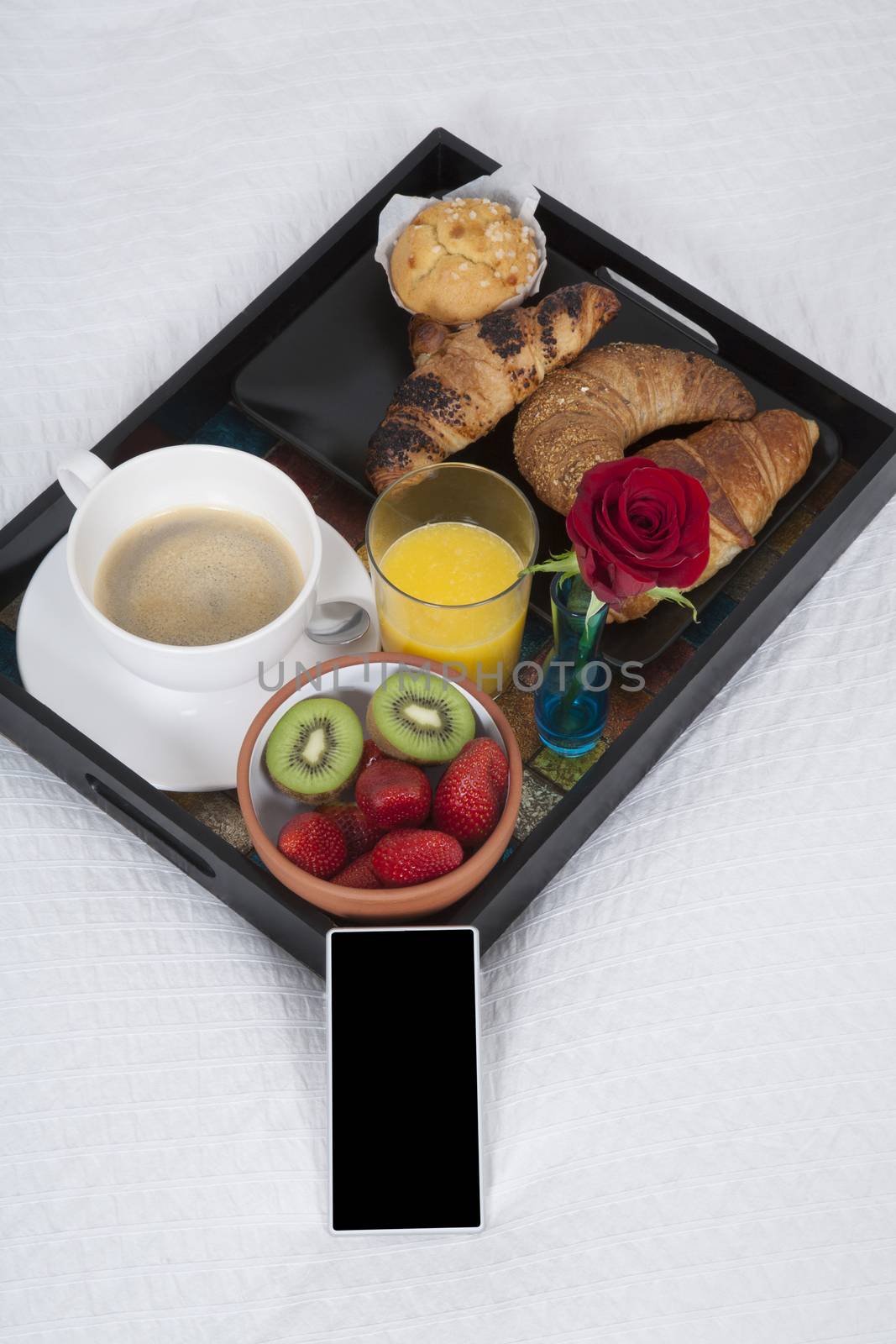 white quilt bed breakfast black tray croissants orange juice strawberry kiwi cupcake red rose flower and smartphone blank screen