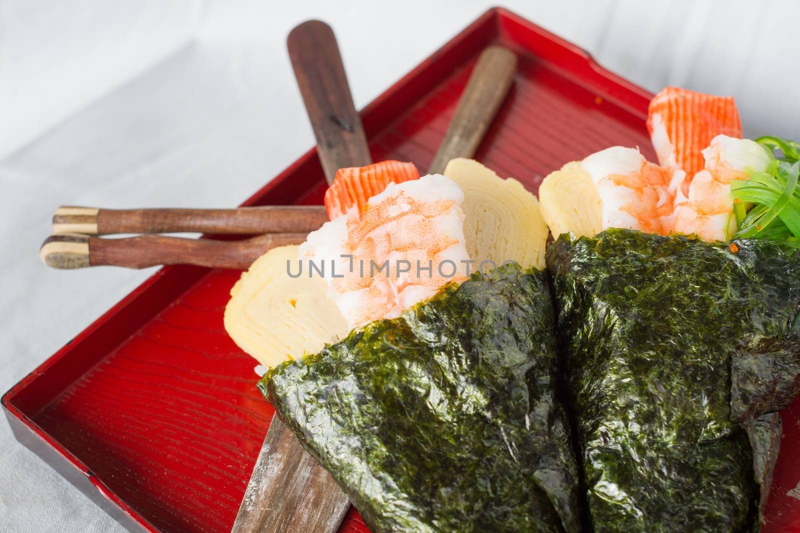 close-up freshly handmade Temaki sushi cone on red rectangular dish with chopsticks and spatula, shrimp focused.