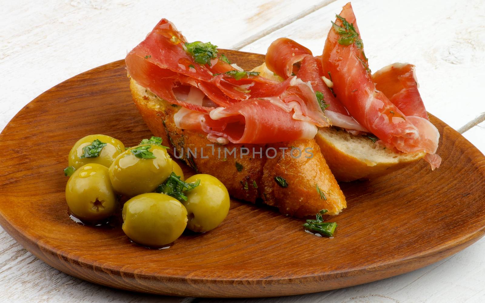 Gourmet Tapas with Smoked Jamon, Garlic Bread and Green Olives on Wooden Plate closeup on White Wooden background