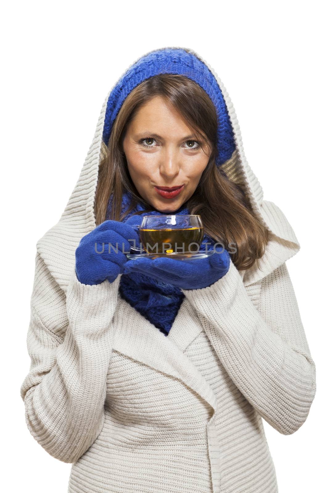 Fashionable young woman in a blue knitted winter ensemble and cowl neck jersey sipping a cup of hot tea with a smile in an effort to keep warm, isolated on white