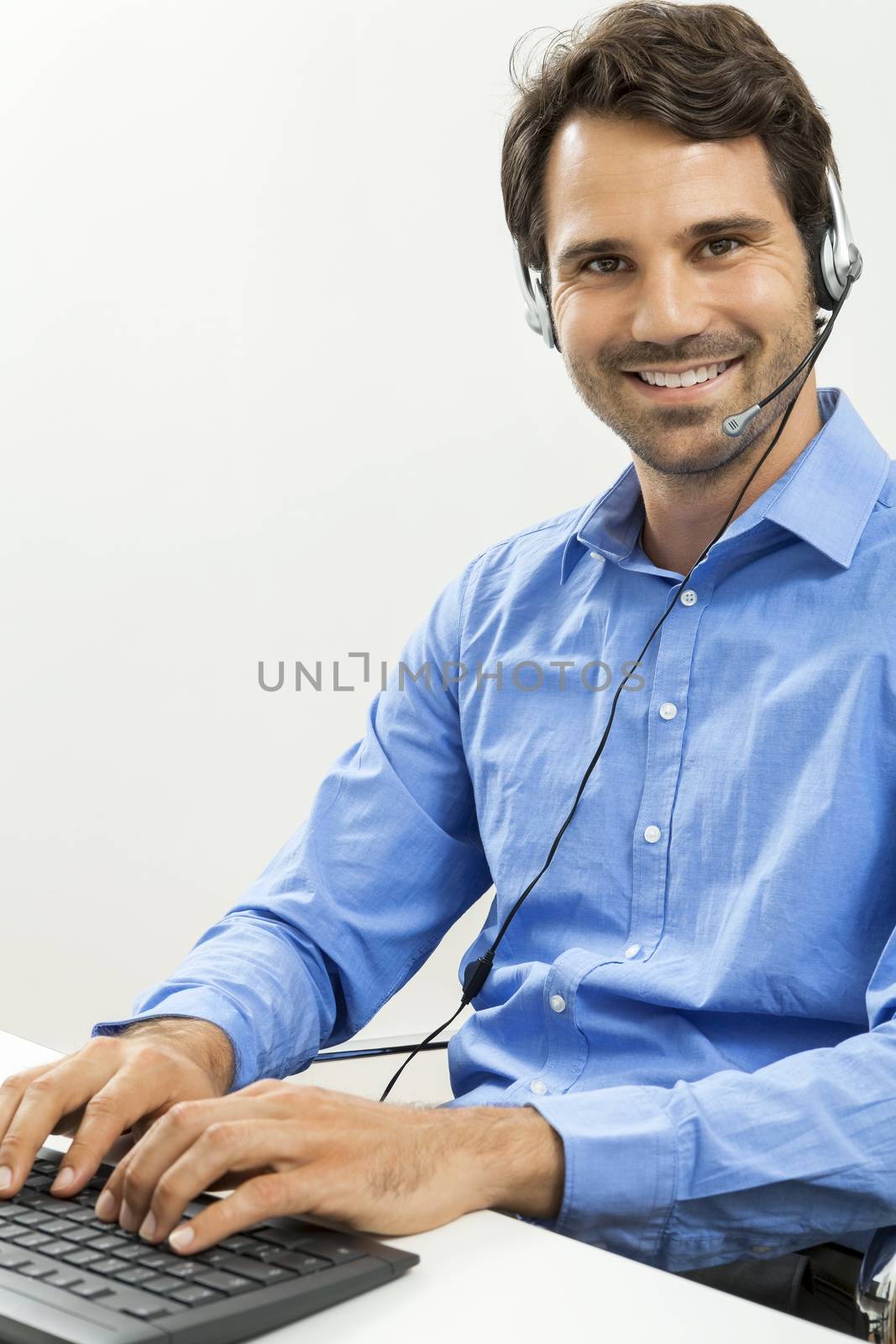 Attractive unshaven young man wearing a headset offering online chat and support on a client services of help desk as he types in information on his computer