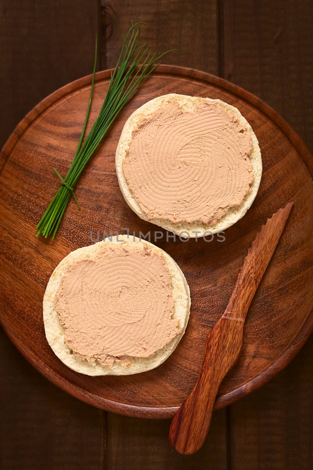 Overhead shot of liverwurst spread on bun with chives and knife on wooden plate photographed with natural light