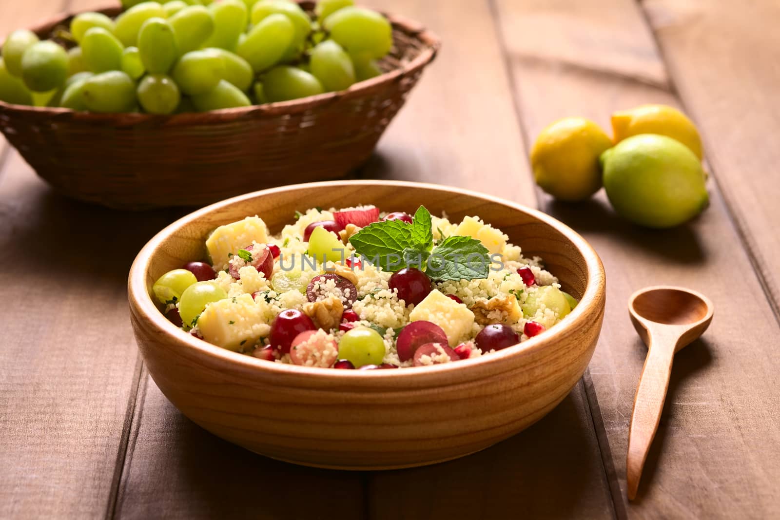 Vegetarian couscous salad with grapes, pomegranate, walnuts, cheese, lime and mint in wooden bowl photographed with natural light (Selective Focus, Focus on the mint leaf on the dish) 