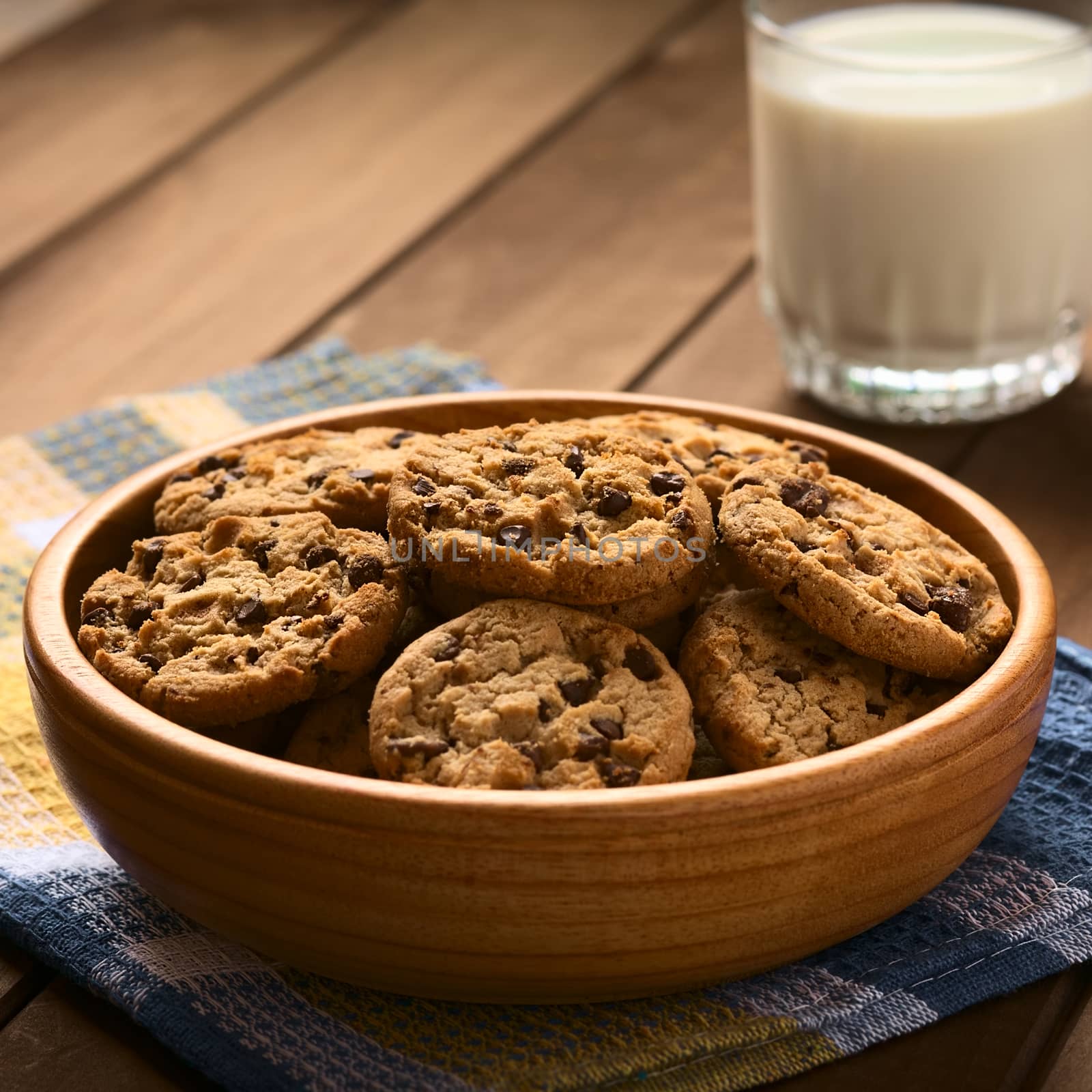 Chocolate chip cookies in wooden bowl with a glass of cold milk in the back, photographed on cloth on wood with natural light (Selective Focus, Focus in the middle of the cookies)