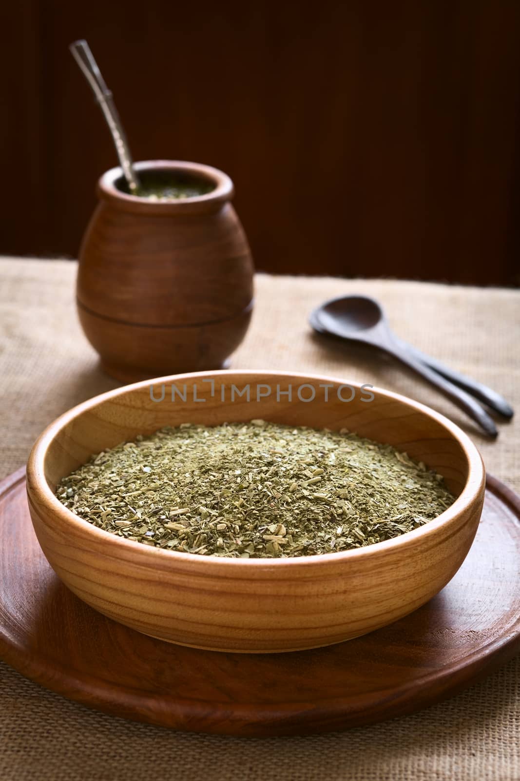 South American yerba mate (mate tea) dried leaves in wooden bowl with a wooden mate cup and strainer (bombilla) in the back, photographed with natural light. Mate is the national infusion of Argentina. (Selective Focus, Focus one third into the dried tea) 