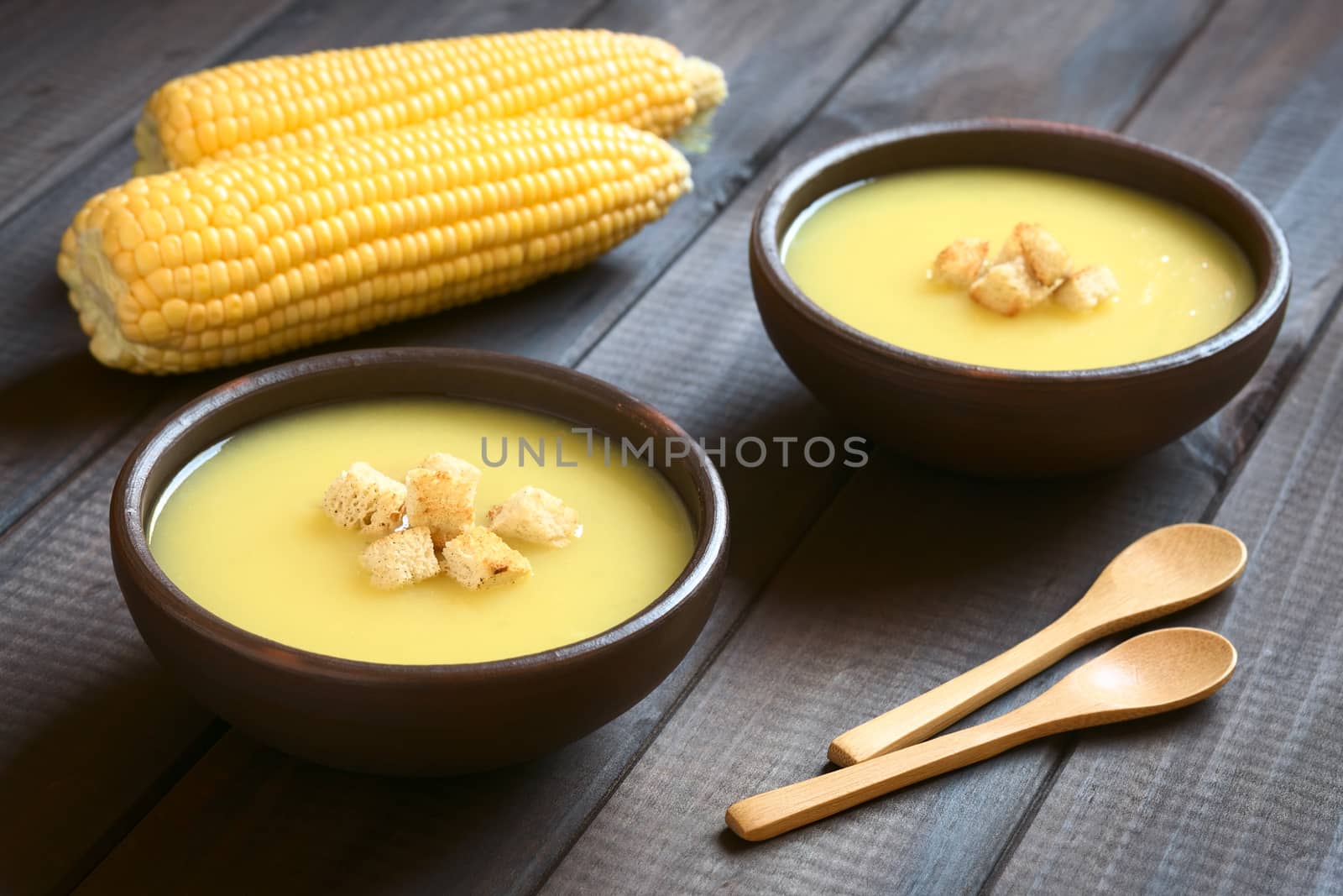 Two bowls of cream of corn soup with croutons on top, photographed on dark wood with natural light (Selective Focus, Focus on the front of the croutons on the first soup)