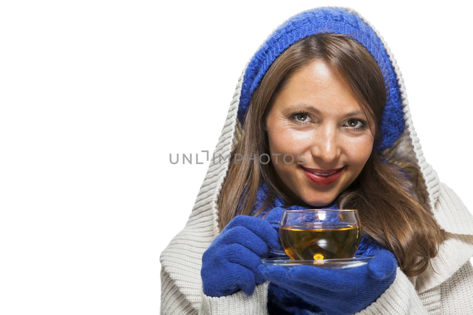 Fashionable young woman in a blue knitted winter ensemble and cowl neck jersey sipping a cup of hot tea with a smile in an effort to keep warm, isolated on white