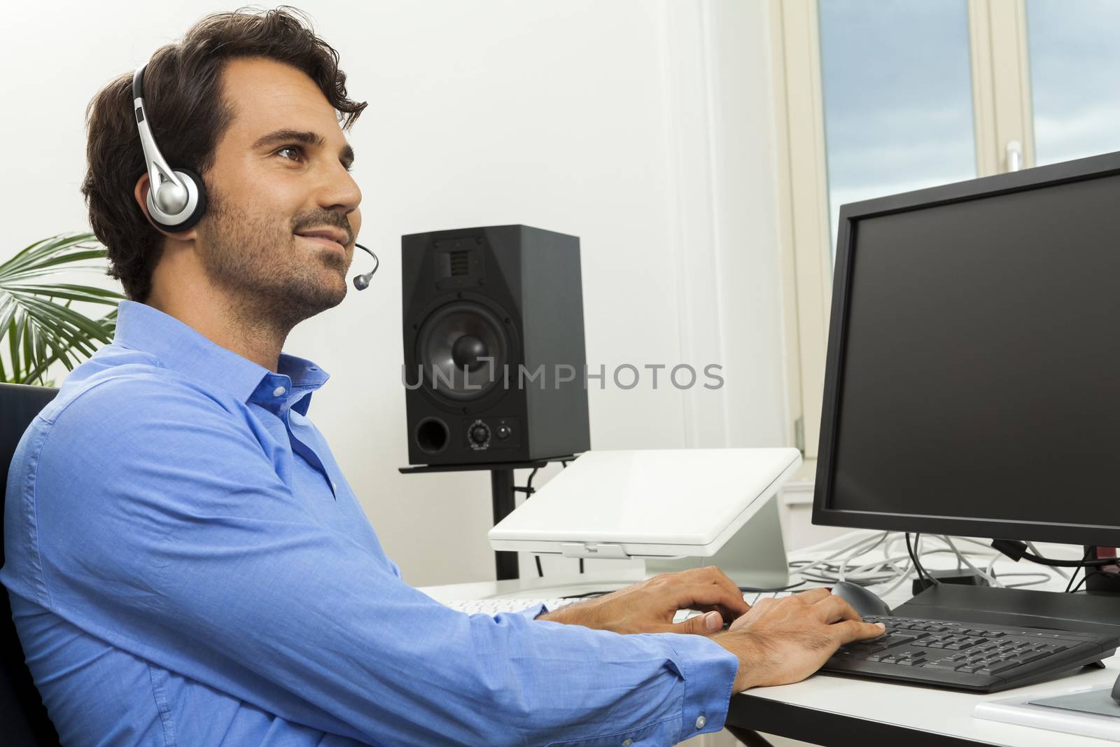 Attractive unshaven young man wearing a headset offering online chat and support on a client services of help desk as he types in information on his computer