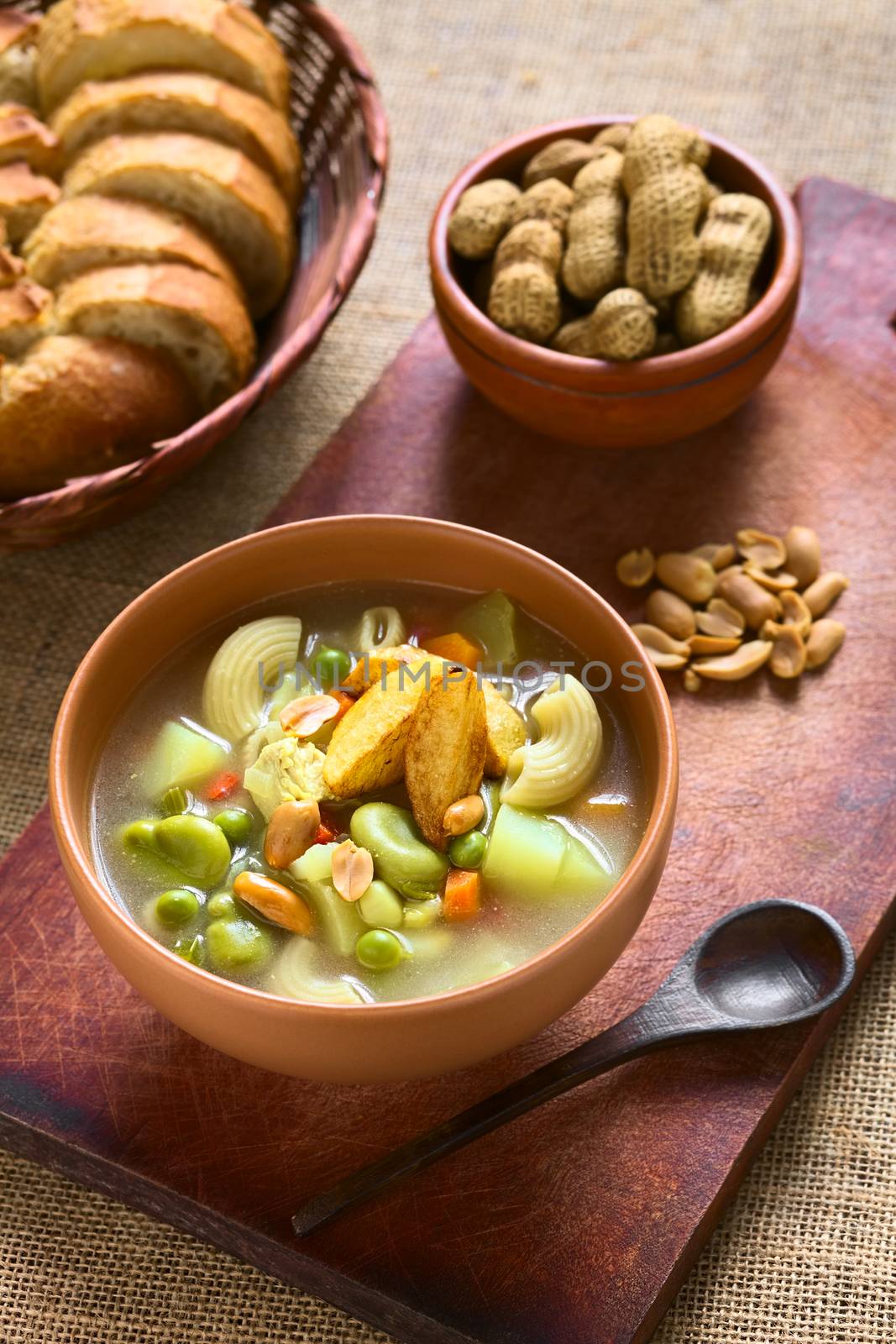 Bowl of traditional Bolivian Sopa de Mani (peanut soup) made of meat, pasta, vegetables (pea, carrot, potato, broad bean, pepper, corn) and ground peanut, traditionally served with fried potatoes, photographed with natural light (Selective Focus, Focus in the middle of the soup)