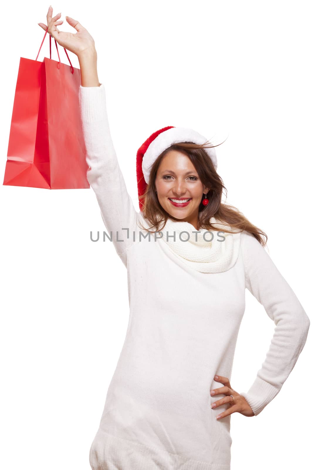 Happy vivacious Christmas shopper wearing a red Santa hat holding up a colorful red shopping bag with a beautiful beaming smile, isolated on white with copyspace