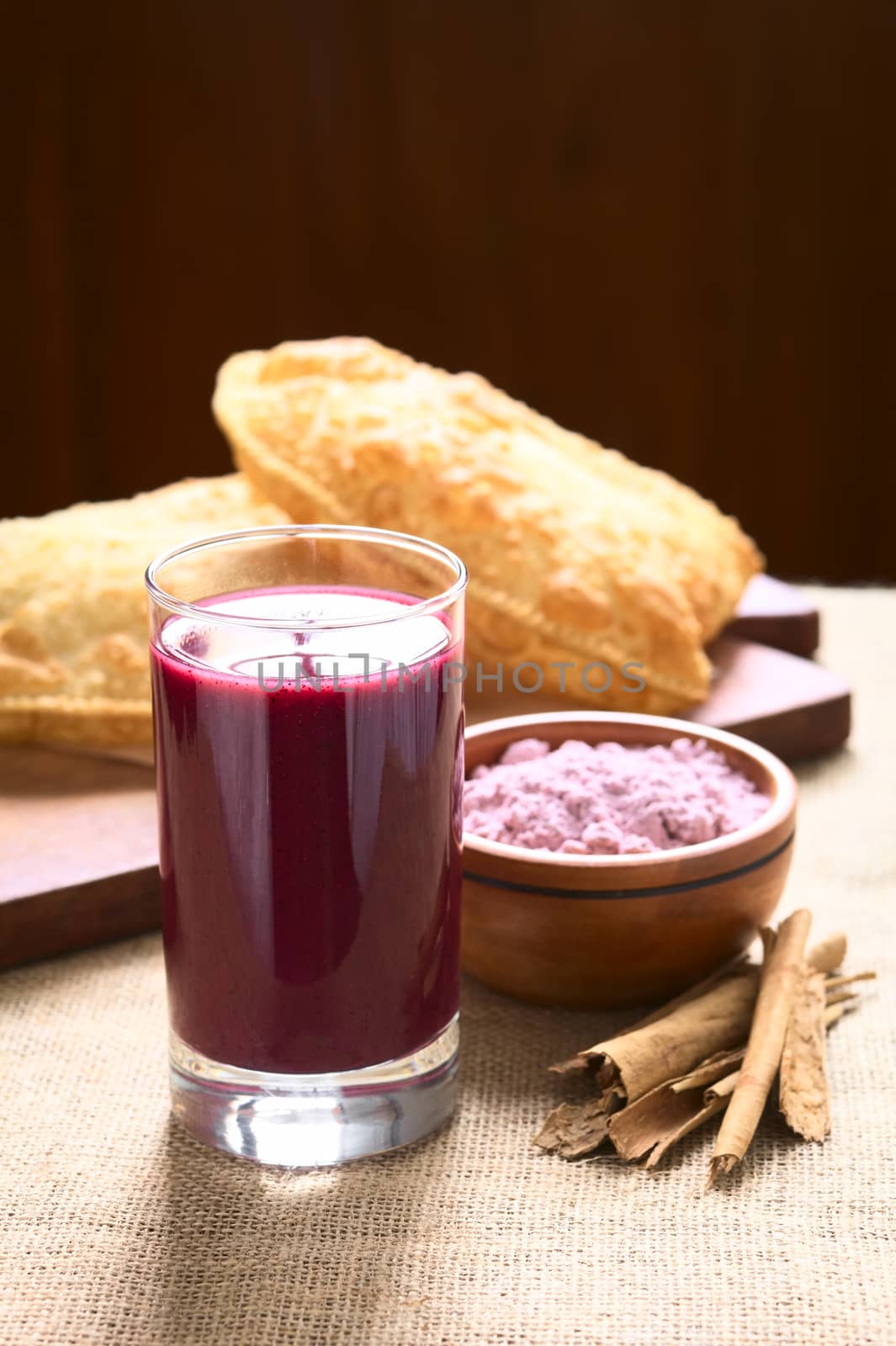 Traditional Bolivian Api, a purple corn beverage, with pastel (deep-fried pastry filled with cheese) in the back, photographed with natural light (Selective Focus, Focus on the front of the drink)