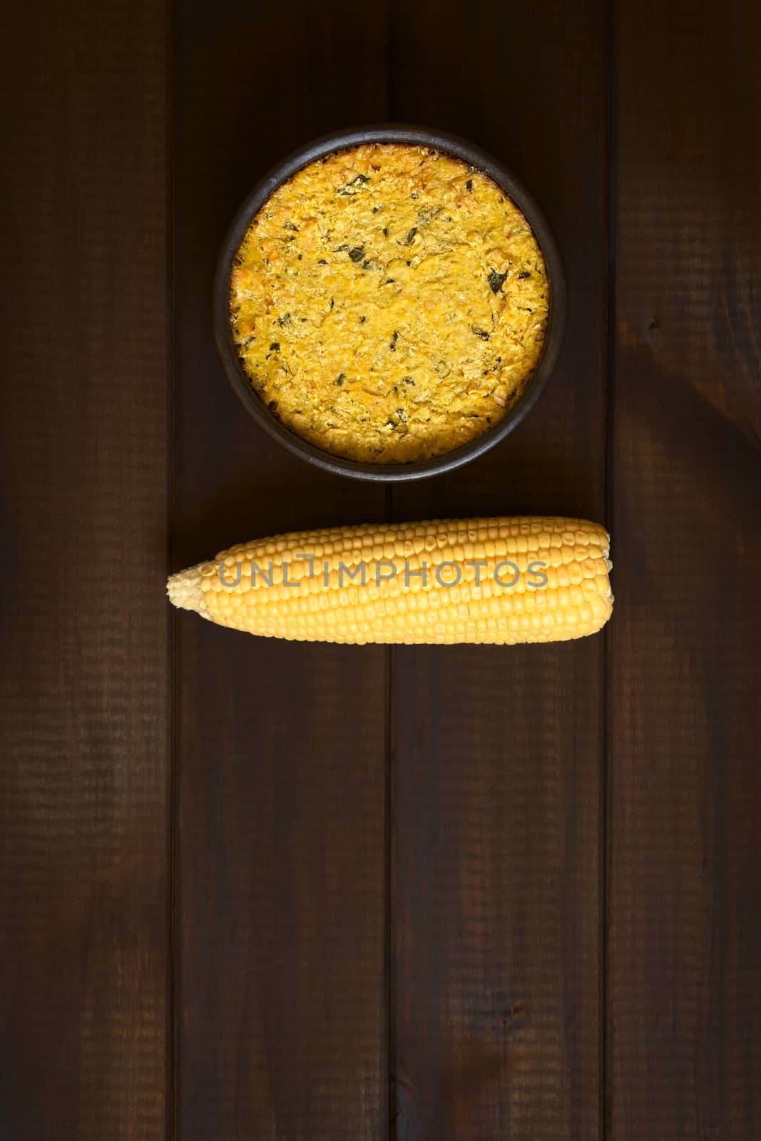 Overhead shot of traditional Chilean corn pie called Pastel de Choclo served in bowl, photographed on dark wood  with natural light. Below the corn-basil mix is ground meat, olive, boiled egg, raisins and pieces of chicken.   