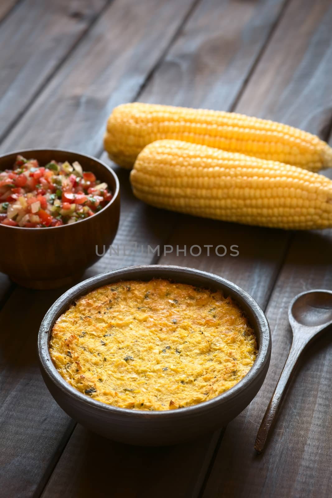 Bowl of traditional Chilean corn pie called Pastel de Choclo served with Pebre sauce and corn cobs in the back, photographed on dark wood  with natural light. Below the corn-basil mix is ground meat, olive, boiled egg, raisins and pieces of chicken. (Selective Focus, Focus in the middle of the dish)