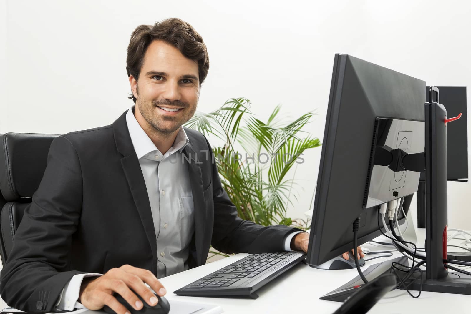 Stylish businessman in a suit sitting at his desk in the office chatting on the phone with a view of his blank computer monitor