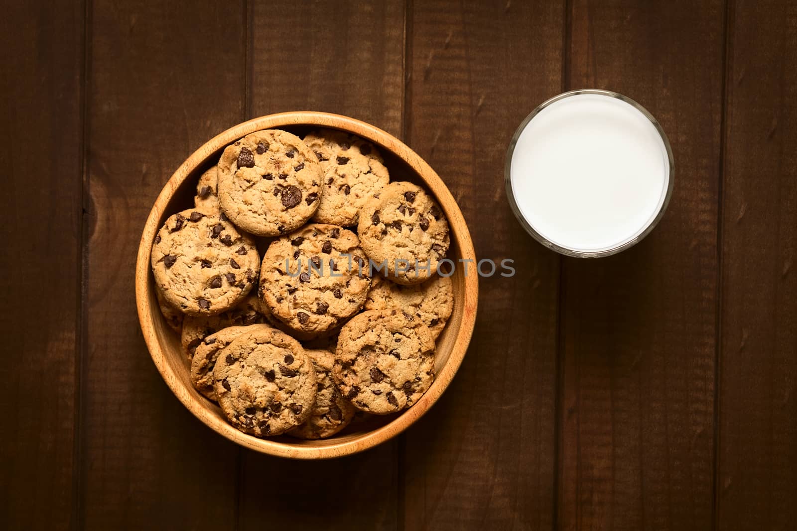 Overhead shot of chocolate chip cookies in wooden bowl with a glass of cold milk on the side photographed on wood with natural light
