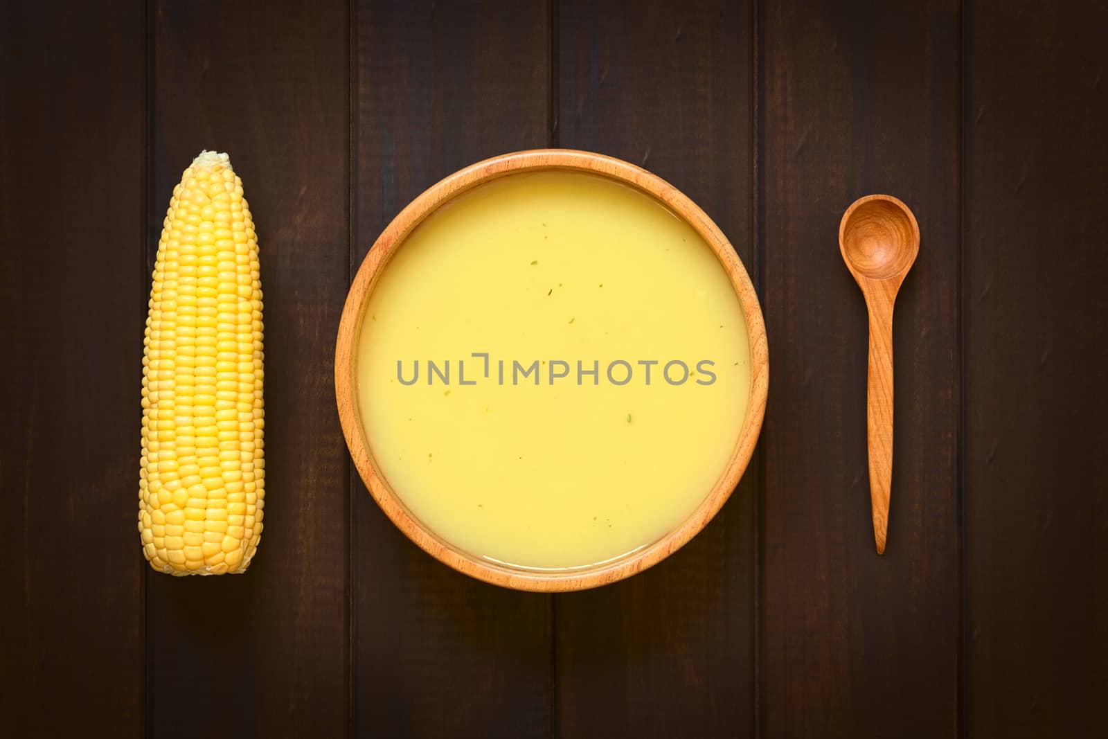 Overhead shot of cream of corn soup in wooden bowl with corn cob and wooden spoon on the side, photographed on dark wood with natural light