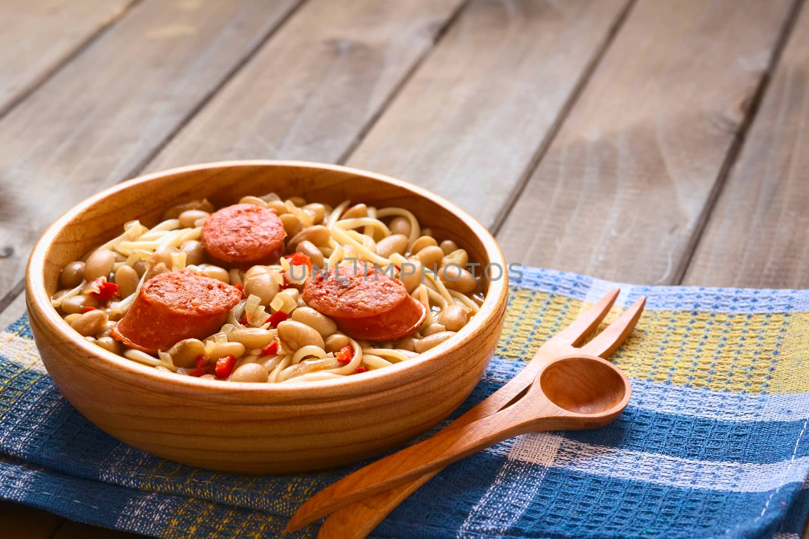 Traditional Chilean dish called Porotos con Riendas (English: beans with reins), made of cooked beans, linguine (flat spaghetti) and served with fried sausage, photographed on dark wood with natural light (Selective Focus, Focus one third into the dish)
