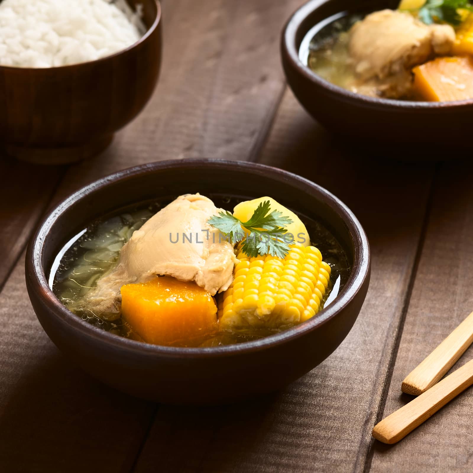 Traditional Chilean Cazuela de Pollo (or Cazuela de Ave) soup made of chicken, sweetcorn, pumpkin and potato, garnished with coriander leaf served in bowl with rice in the back (which is usually eaten with the soup), photographed on wood with natural light (Selective Focus, Focus in the middle of the soup)