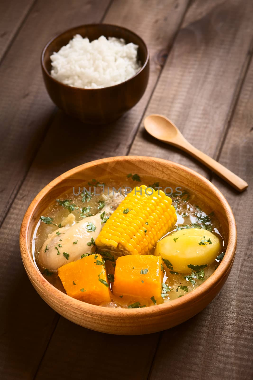 Traditional Chilean Cazuela de Pollo (or Cazuela de Ave) soup made of chicken, sweetcorn, pumpkin and potato, seasoned with fresh coriander served in wooden bowl with rice in the back (which is usually eaten with the soup), photographed on wood with natural light (Selective Focus, Focus one third into the soup)