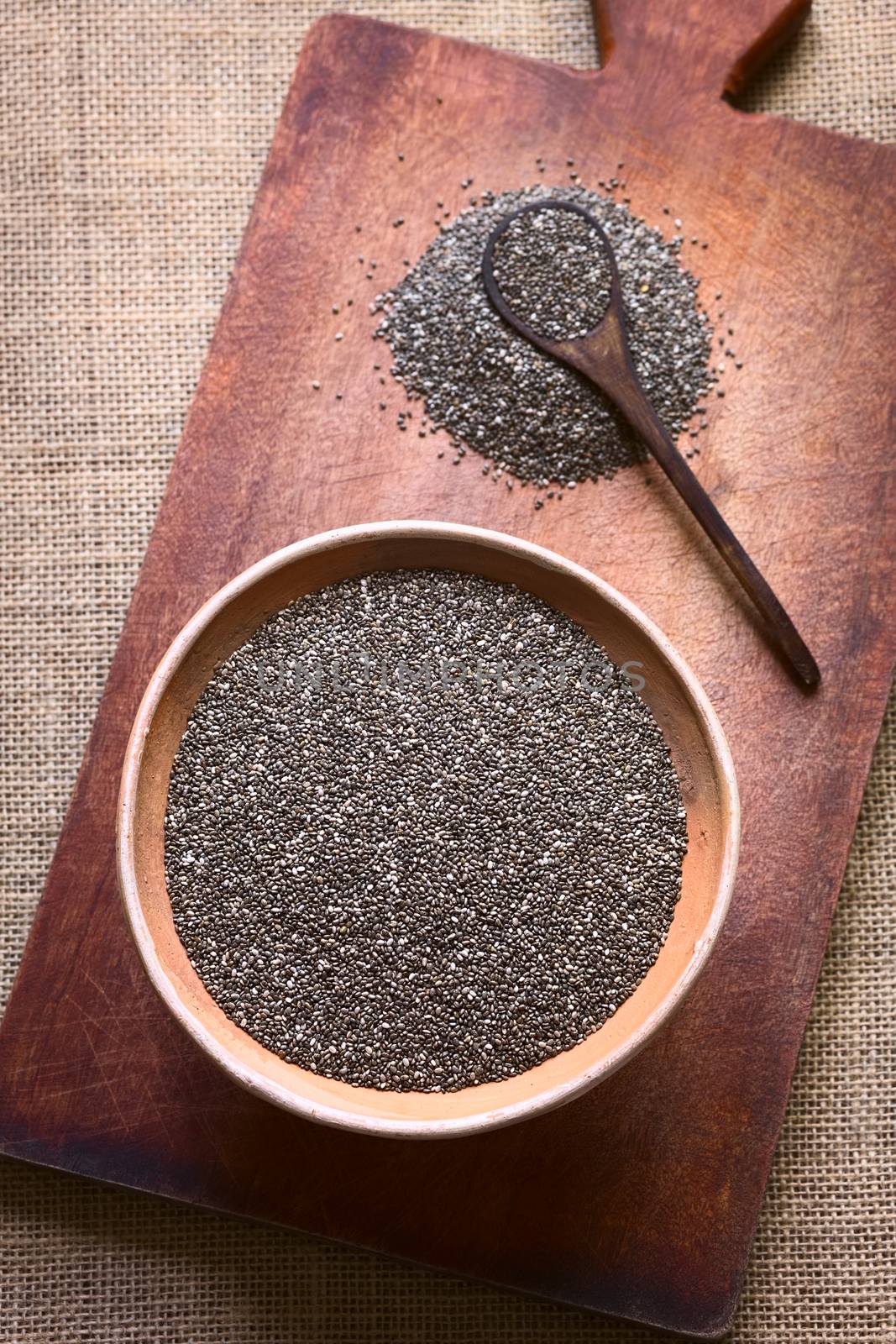 Overhead shot of chia seeds (lat. Salvia hispanica) in clay bowl photographed on wooden board with natural light. Chia seeds are considered a superfood containing proteins, omega fats, minerals and antioxidants (Selective Focus, Focus on the seeds in the bowl)