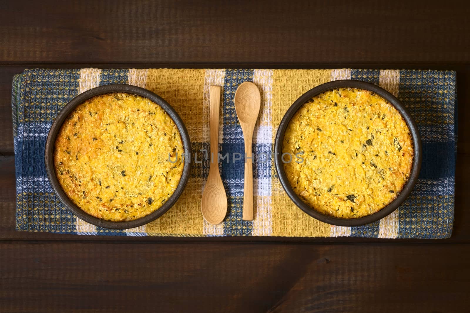 Overhead shot of traditional Chilean corn pie called Pastel de Choclo served in bowls, photographed on cloth on dark wood  with natural light. Below the corn-basil mix is ground meat, olive, boiled egg, raisins and pieces of chicken.   