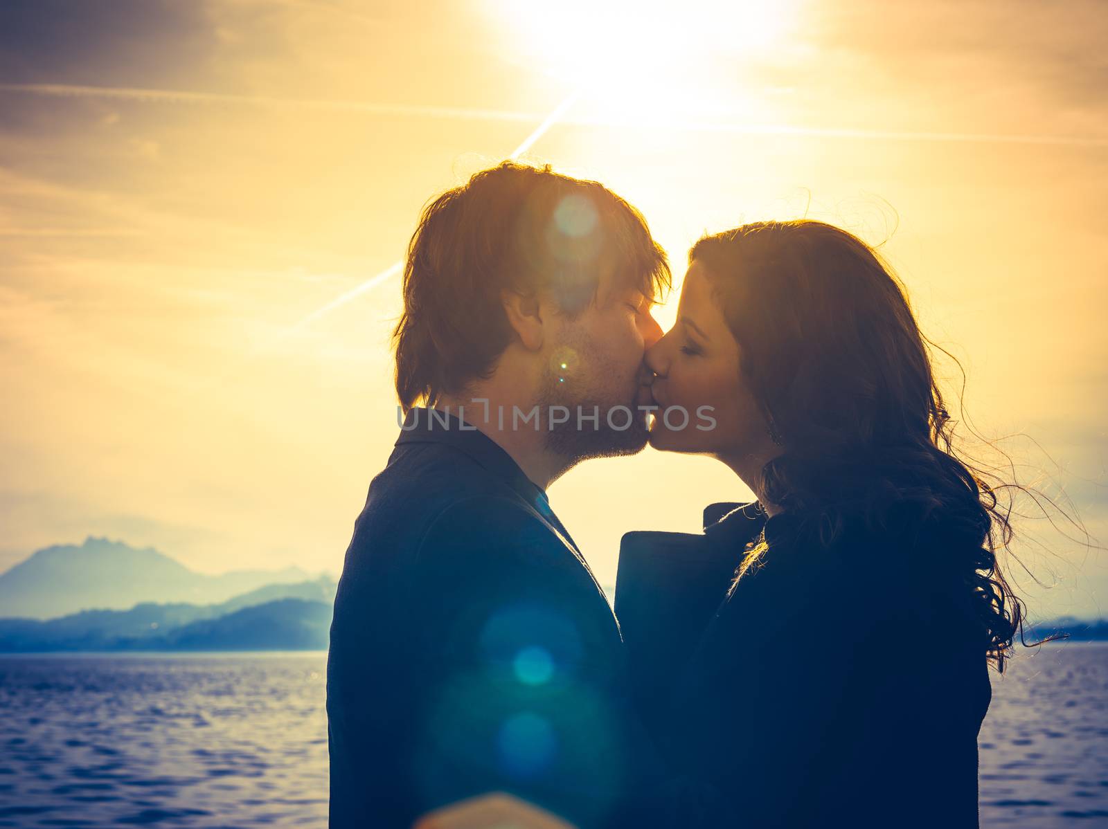 Photo of young adults by the water hugging and kissing in the late afternoon as the sun lowers in the sky. Heavily filtered for more romantic effect. Real lens flare visible.