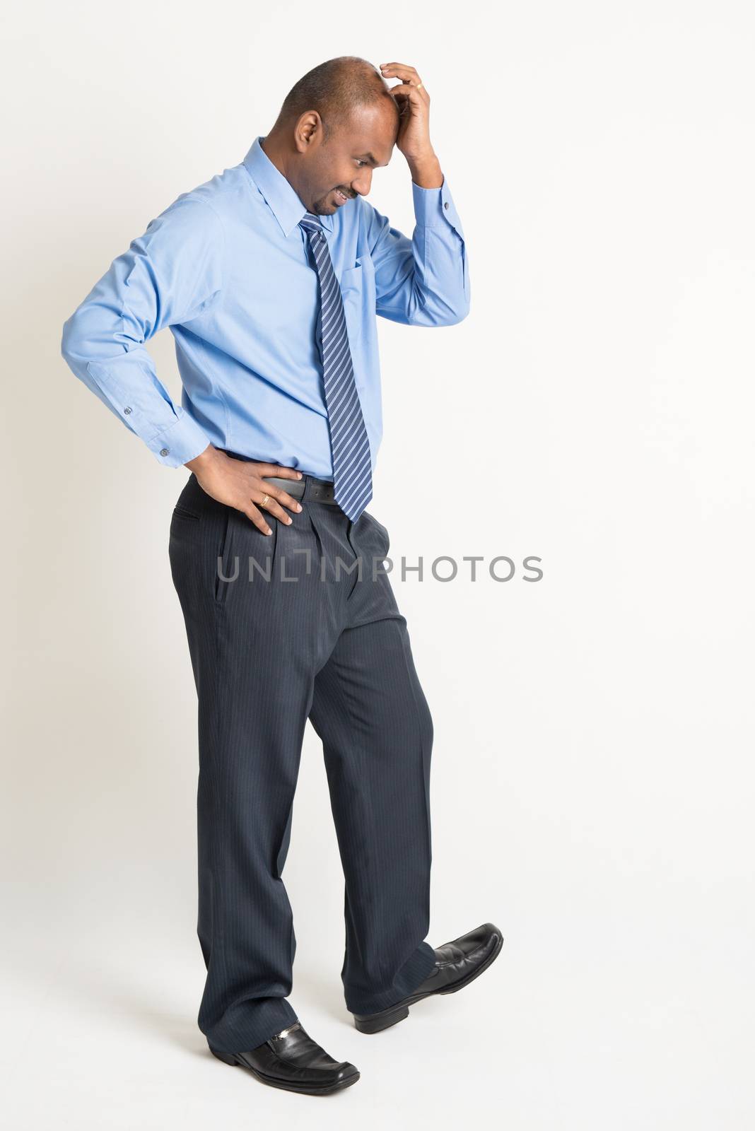 Full length Indian businessman with worried expression standing on plain background with shadow.