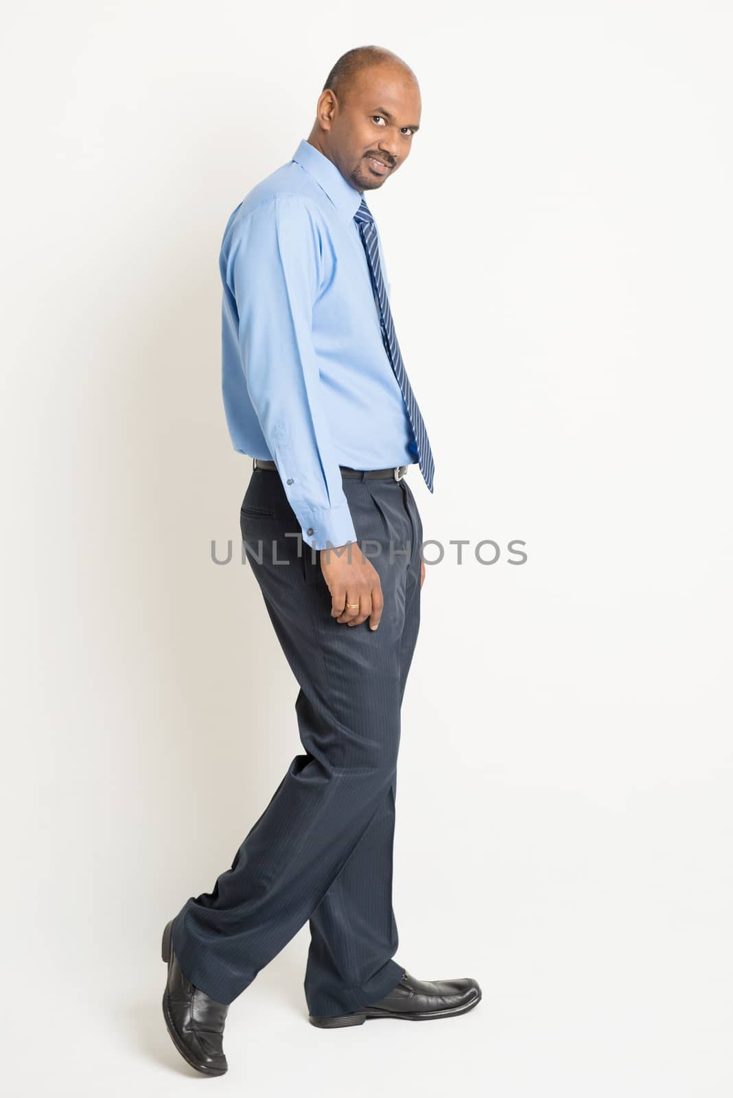 Side view Indian businessman walking and turn back head looking at camera, on plain background.