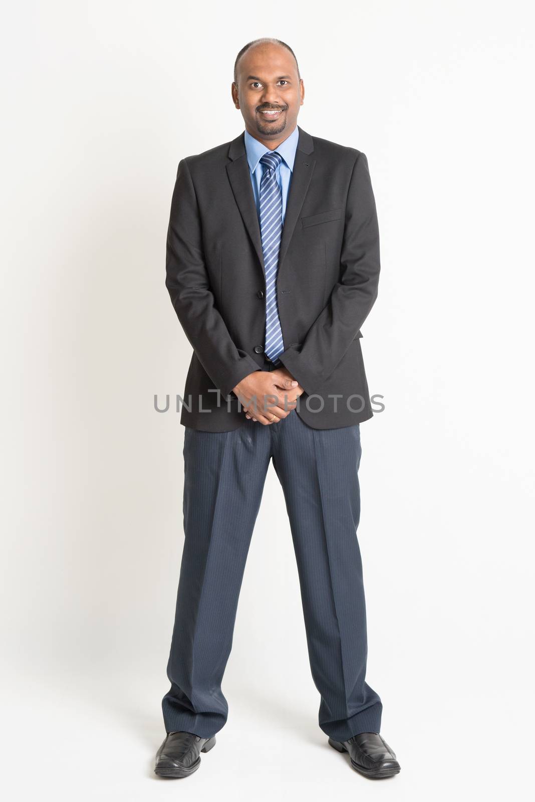 Full length friendly Indian businessman in formal suit looking at camera, standing on plain background.
