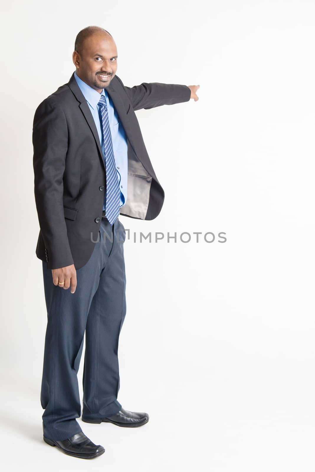 Full body Indian businessman pointing away to copy space, on plain background.