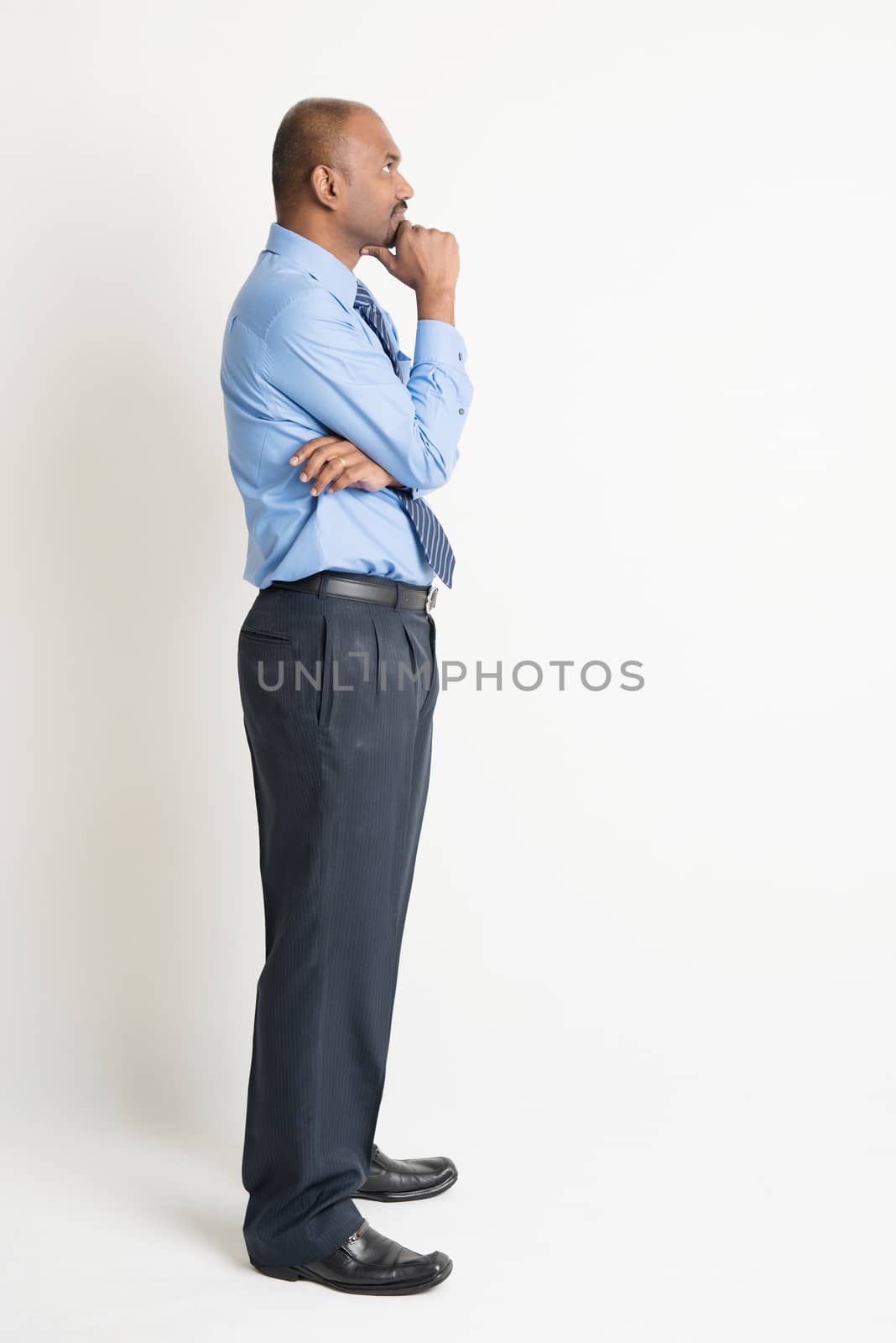 Profile view of full body Indian businessman hand on chin looking up at blank copy space, standing on plain background with shadow