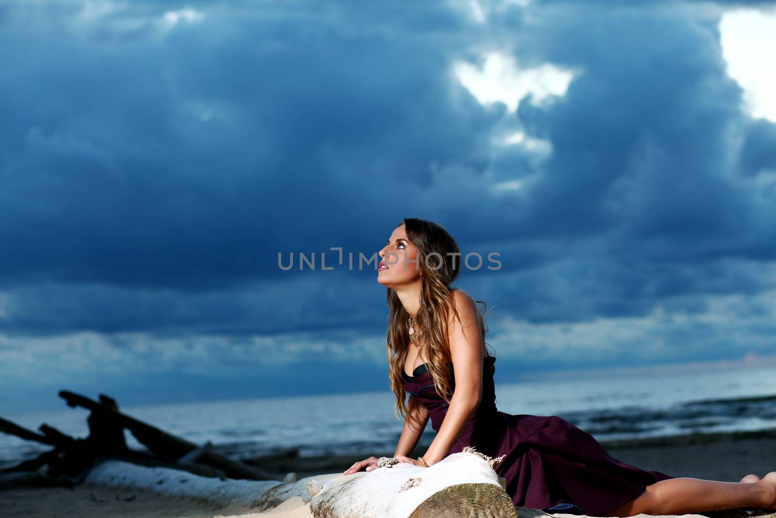 Beautiful girl with brown hair who is wearing a dark purple dress is posing at a beach over a dark sky background