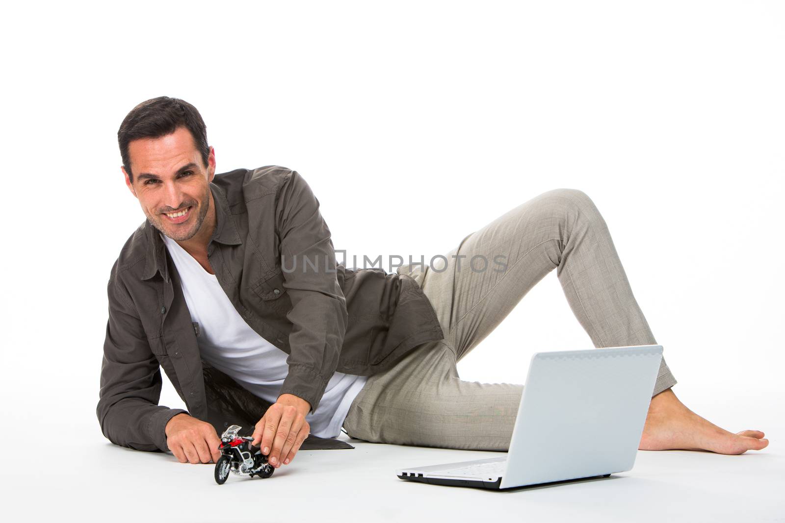 Man laying on the floor, smiling at camera playing with a motorbike scale model and laptop next to him